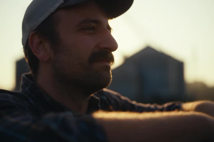 A farmer with a mustache and hat relaxes and smiles while enjoying the golden hour sunset on his farm