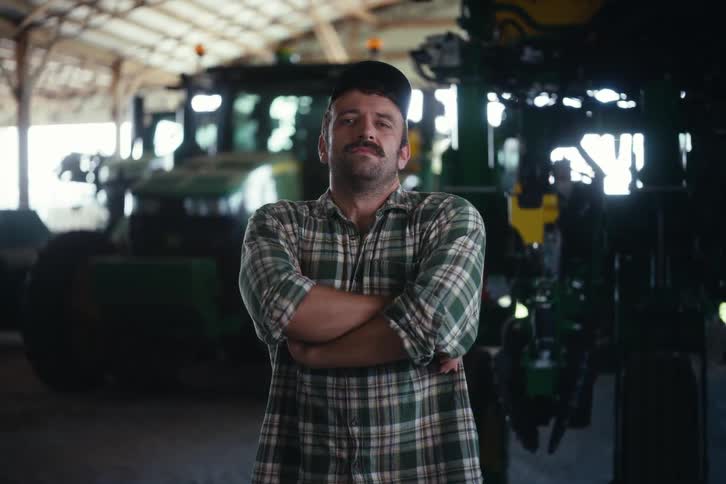 A proud mustachioed farmer looks up and crosses his arms, standing confidently in a barn in front of his tractors
