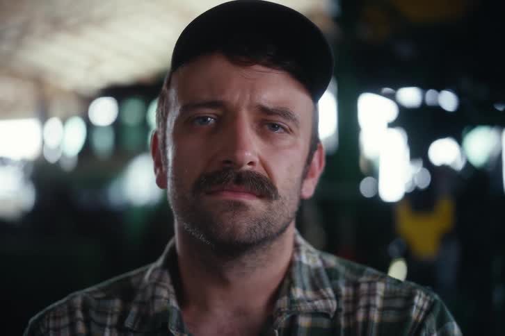 Close-up portrait of a serious farmer with a mustache and cap, looking up with a solemn expression inside a barn