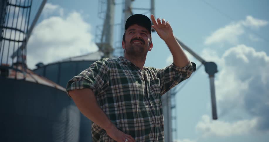 A proud and happy farmer in a plaid shirt and cap surveys his land in slow motion in front of massive grain silos