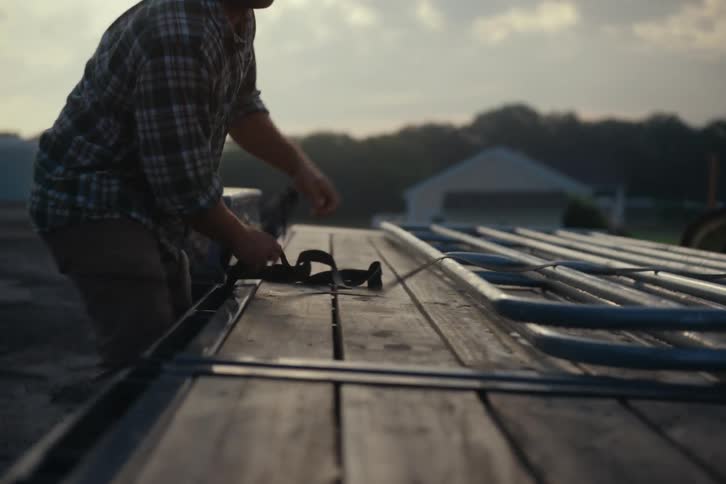 A male farmhand wearing a plaid shirt and a baseball cap secures metal gates onto a flatbed trailer using a ratchet strap at sunset