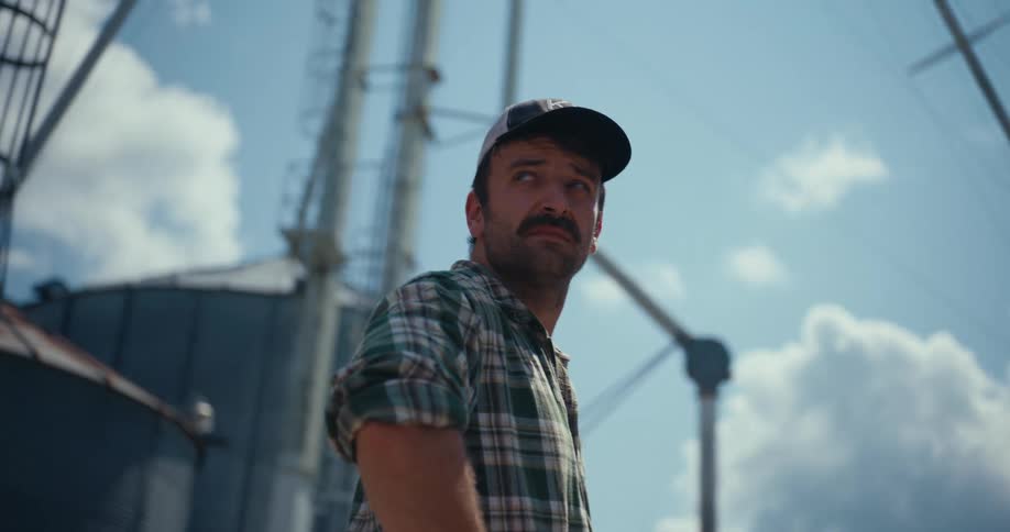 A thoughtful farmer with a mustache stands between large grain silos and looks up at the sky on a sunny day
