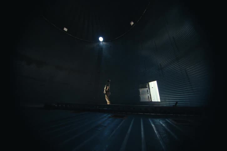 A solitary man stands inside a vast, dark, empty grain silo, illuminated by a single light from above