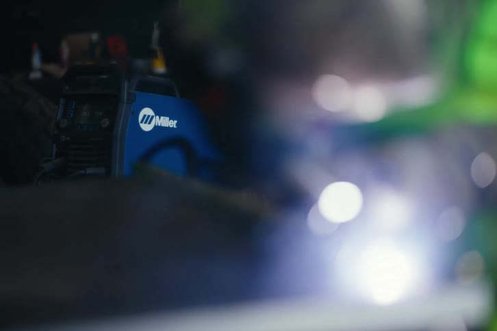 A Miller welding machine sits in focus in a dark workshop while bright sparks fly from out-of-focus welding in the foreground