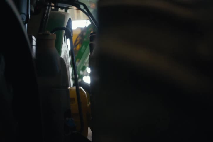 A farmer in a plaid shirt climbs down from his tractor in a dimly lit machine shed and walks towards the light