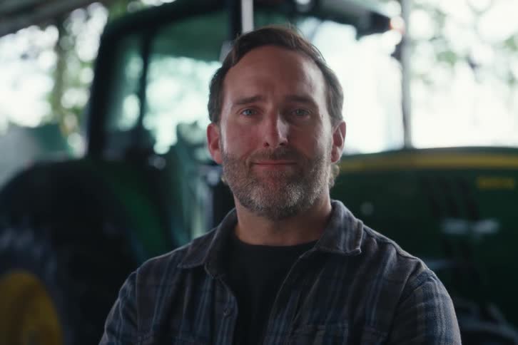 A hardworking farmer with a beard and plaid shirt smiles proudly in front of a green tractor in a barn