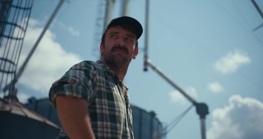 A thoughtful farmer in a plaid shirt turns to look up at the towering grain silos on a sunny day