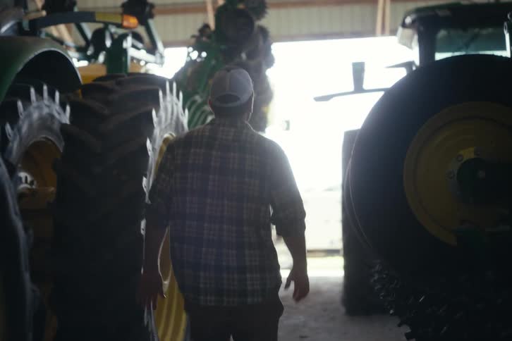 A farmer walks in slow motion through a large barn filled with tractors and other farming equipment