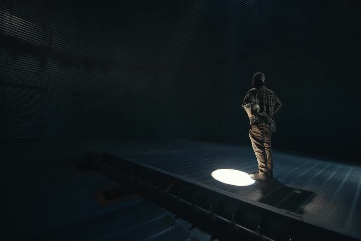 A lone farmer stands under a single spotlight, contemplating the vast, dark, empty space inside a metal grain silo