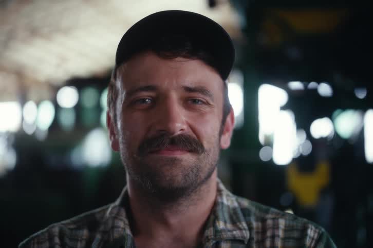 A mustachioed man in a cap and flannel shirt looks up and smiles directly at the camera in a soft-focus workshop setting