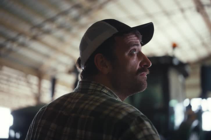 A mustached farmer in a plaid shirt and baseball cap turns and adjusts his hat while standing inside a barn