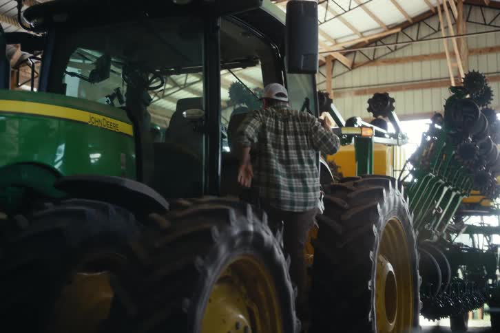 A farmer walks through a barn and climbs into the cab of his green tractor to begin his work day