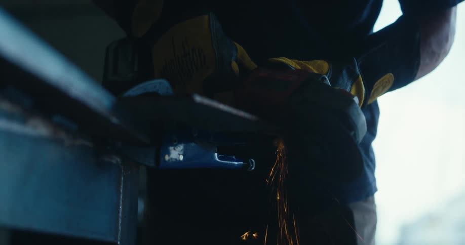 A worker in protective gloves uses an angle grinder on a piece of metal, sending sparks flying in slow motion