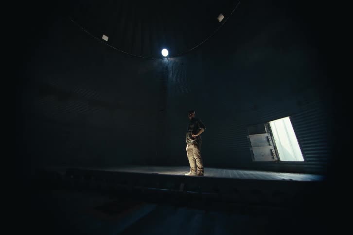 A solitary man stands in a single spotlight inside a vast, empty grain silo as the camera slowly pulls back