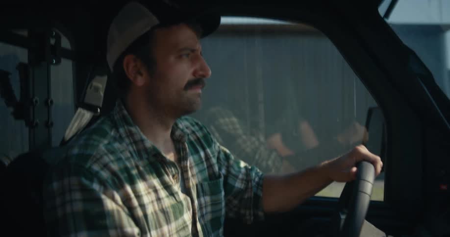 A mustached farmer in a plaid shirt and baseball cap drives a utility vehicle on the farm