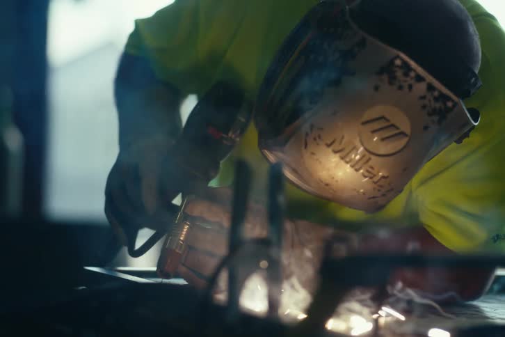 Cinematic slow-motion close-up of a welder in a protective mask joining metal, with bright sparks and smoke filling the air