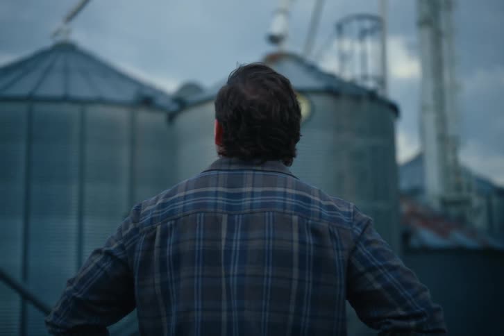 A farmer stands with his back to the camera, looking up at large grain silos on his farm at dusk