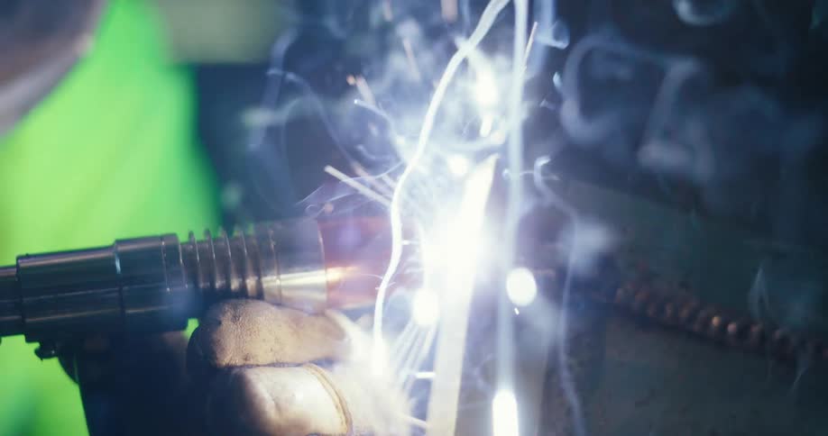 Extreme close-up slow-motion shot of a welder joining two pieces of metal, creating bright sparks and plumes of smoke