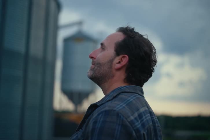 A pensive farmer wearing a plaid shirt looks up with a slight smile while standing in front of large metal grain silos at dusk