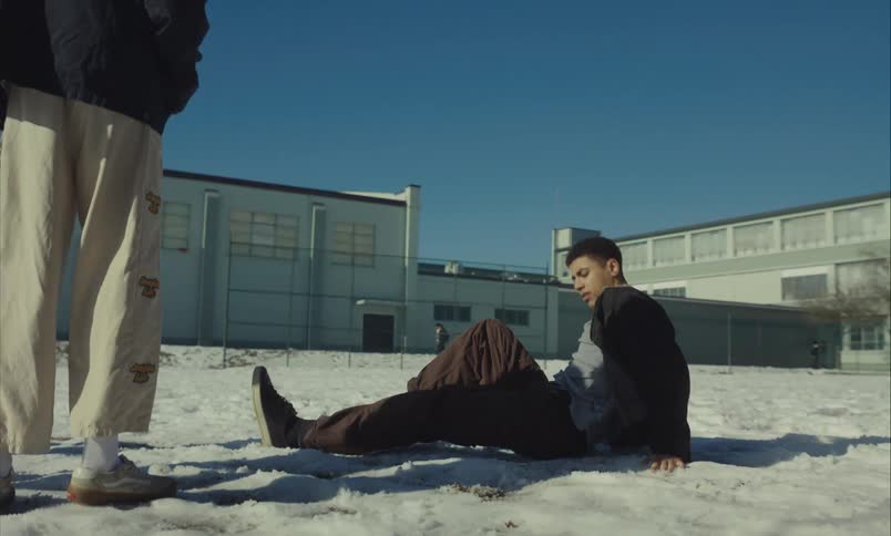 A low-angle shot reveals a teenage boy lying on the snowy ground as two other boys stand over him in a schoolyard on a sunny winter day