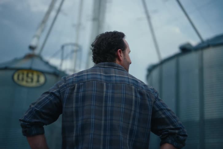 A proud farmer in a flannel shirt stands with his hands on his hips, surveying his large grain silos on a cloudy evening
