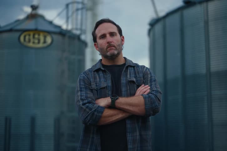 A stoic farmer with his arms crossed looks up and stares intently at the camera while standing in front of two grain silos