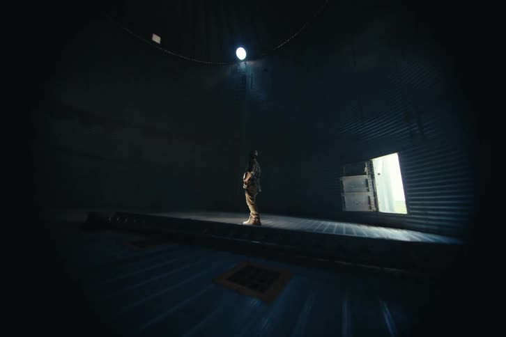 Slow push-in shot of a farmer standing alone in the center of a dark, empty grain silo, looking up at a single light source
