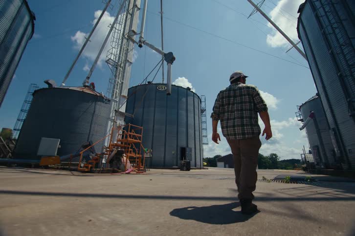 Low angle slow-motion shot of a farmer walking between large grain silos on a bright sunny day
