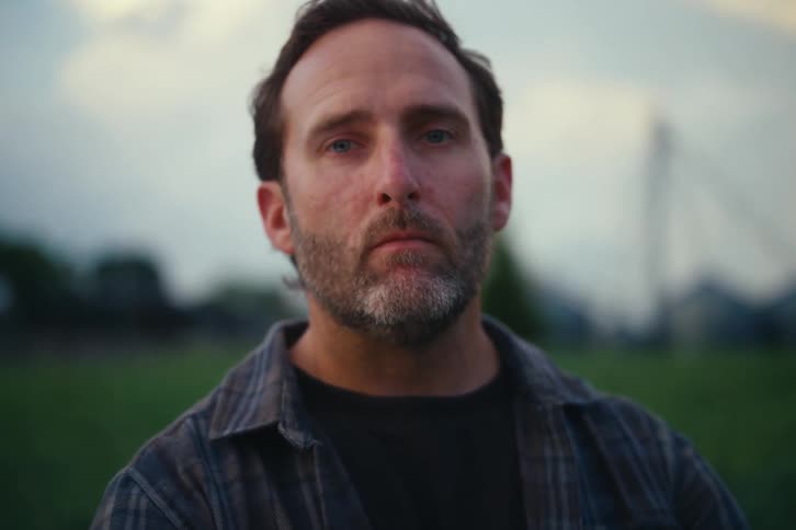 A dramatic close-up portrait of a serious, bearded man looking directly into the camera while standing in a field at dusk