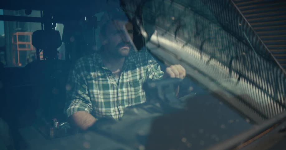 A mustached farmer in a plaid shirt drives a combine harvester on his farm, seen through the reflective windshield