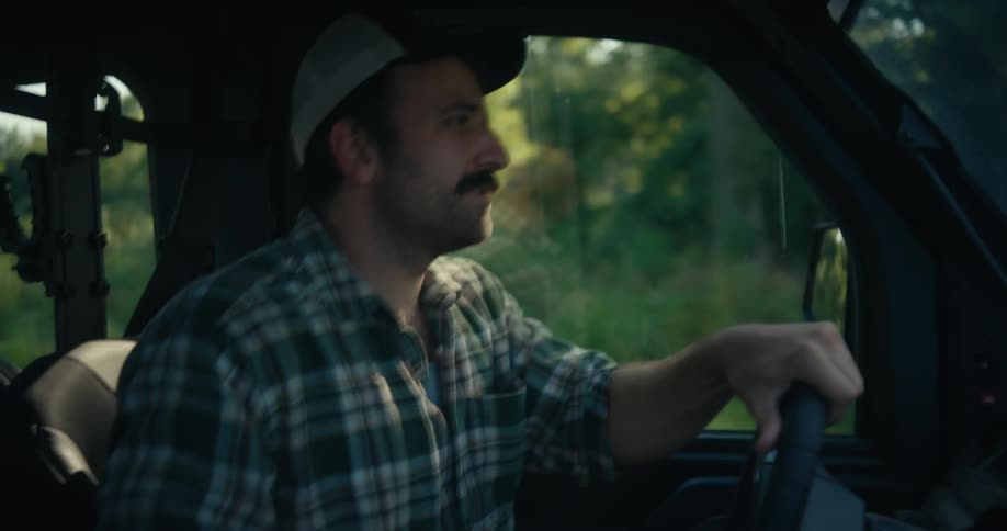 A mustached man in a plaid shirt and baseball cap drives a utility vehicle down a rural road in slow motion
