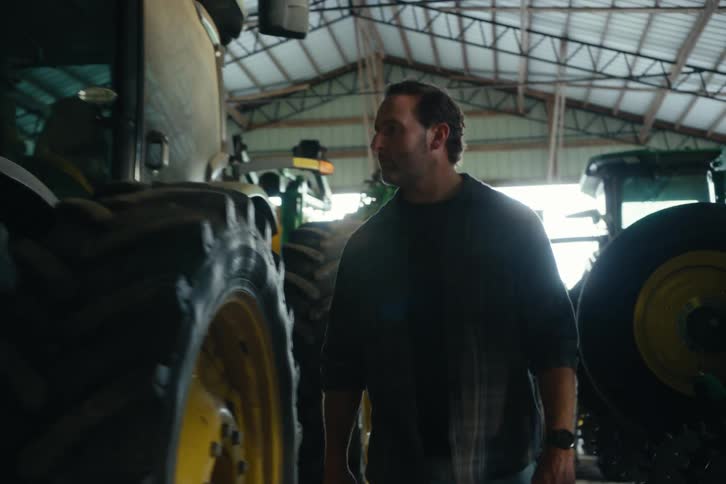 A serious farmer walks in slow motion through a large barn filled with green tractors and heavy farming machinery