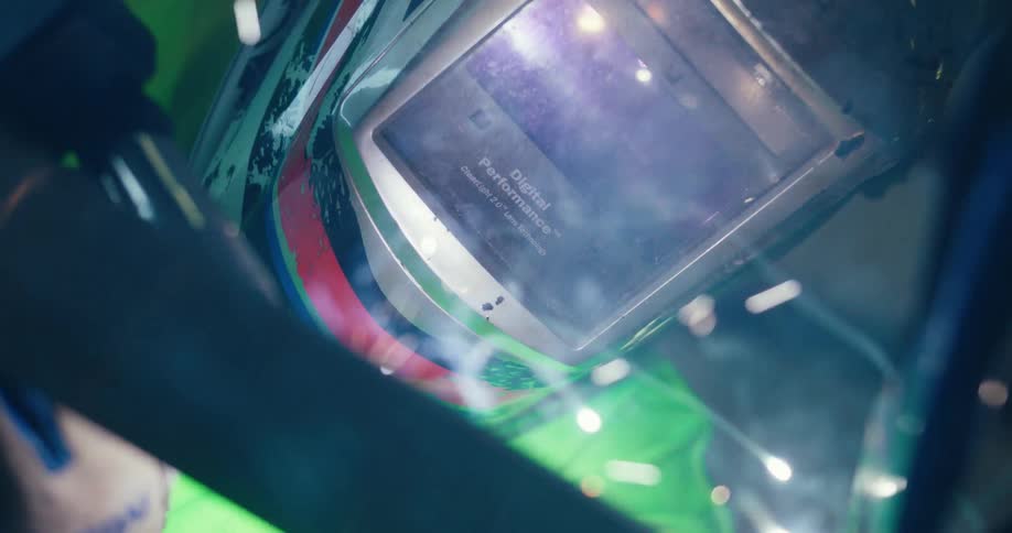A dramatic close-up of a welder's mask reflects the bright light, sparks, and smoke from the welding arc below
