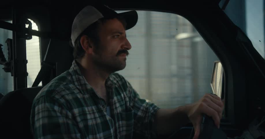 A serious farmer with a mustache wearing a plaid shirt and baseball cap drives a truck through a rural farm landscape passing by silos