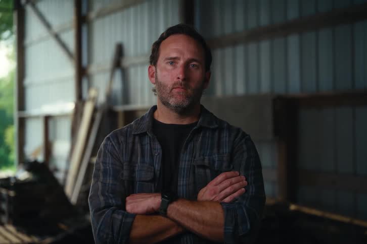 A serious, blue-collar man in a flannel shirt looks up and crosses his arms while standing in a rustic barn