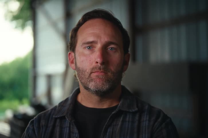 A serious, bearded man in a flannel shirt looks up and stares intently at the camera inside a rustic barn