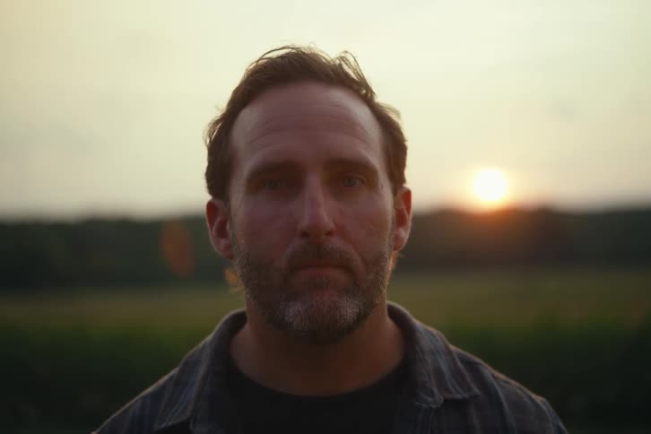 A pensive man with his eyes closed slowly opens them to look directly into the camera with the sunset in the background
