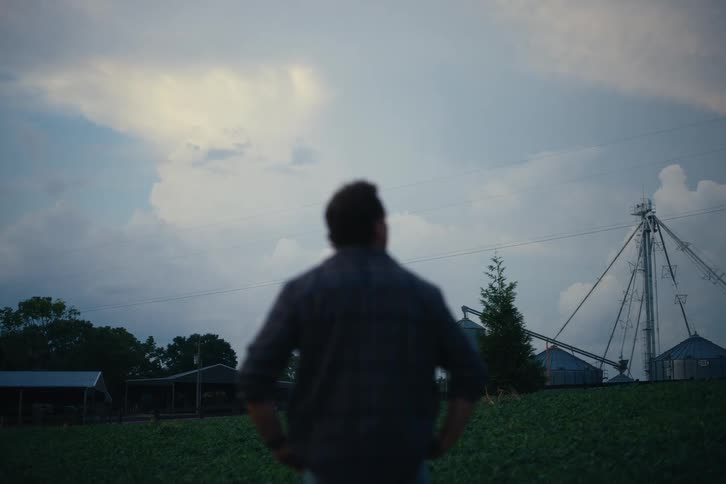 Out-of-focus shot of a farmer standing in a field at dusk, looking out at his grain silos