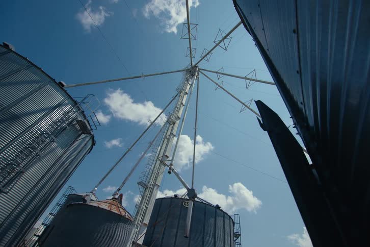 A slow, low-angle tilt-up shot reveals massive grain silos against a blue sky with fluffy clouds on a sunny day
