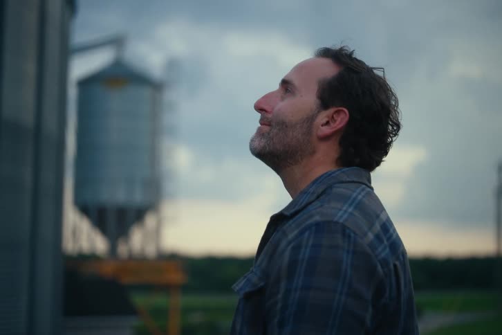 A proud farmer wearing a plaid shirt looks up at his grain silo with a smile on a cloudy evening