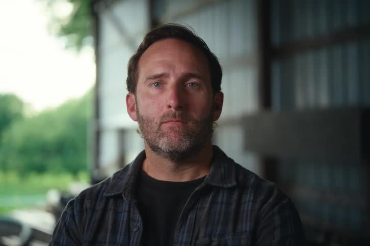 Portrait of a serious, rugged middle-aged man in a flannel shirt turning to look at the camera inside a barn