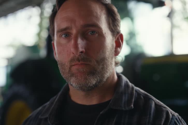 A serious, bearded farmer in a flannel shirt looks up with an intense stare while standing in his barn