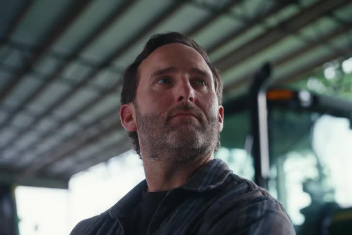 A serious-looking farmer with a beard stands thoughtfully inside a barn next to his tractor
