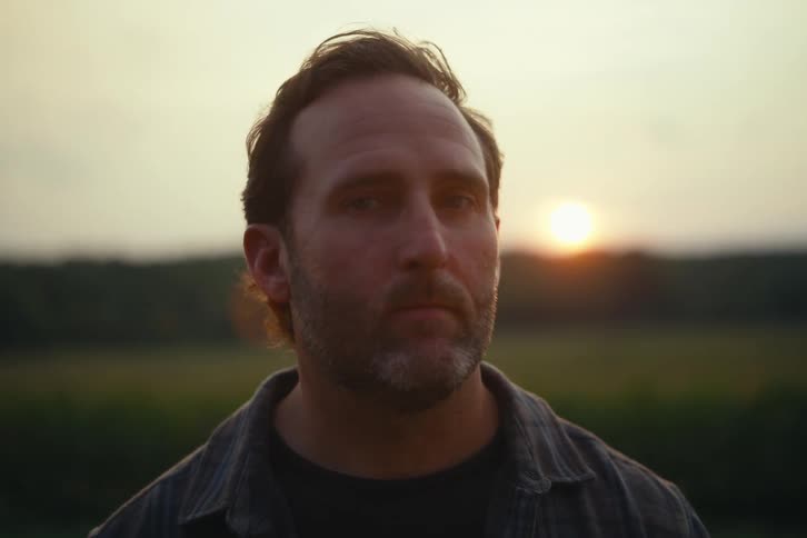 A pensive, bearded man stands in a field and turns to look seriously at the camera during a golden sunset
