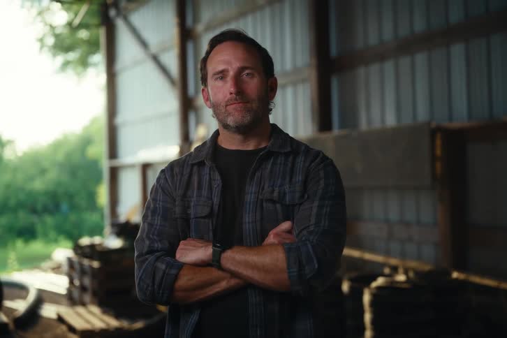 A confident blue-collar man in a plaid shirt stands with his arms crossed in a barn and looks directly at the camera