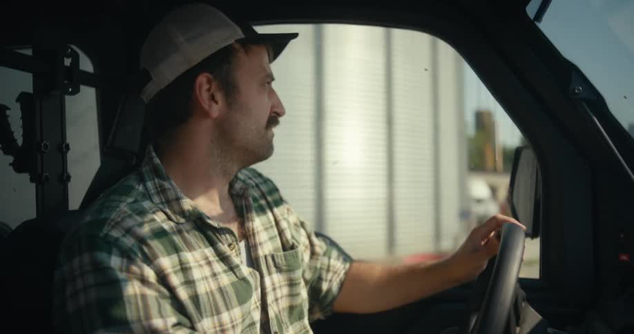 A focused farmer with a mustache drives a utility vehicle around his farm, passing by fields and large grain silos