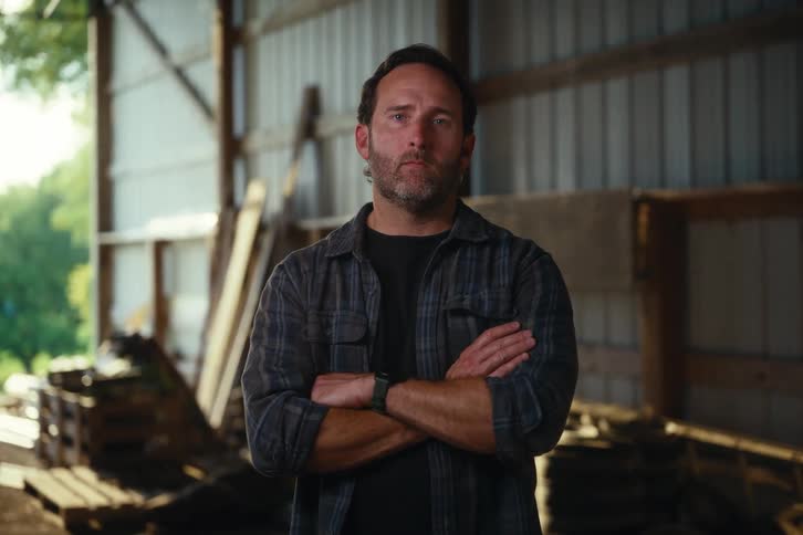 Portrait of a serious man with a beard crossing his arms and looking directly at the camera while standing in a rustic barn