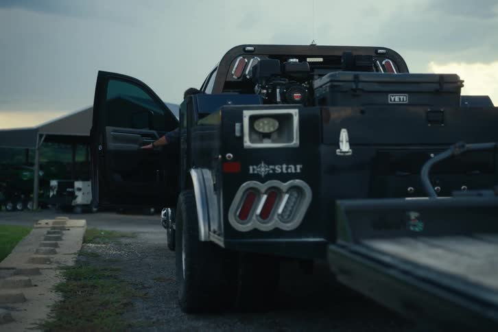 A man in a plaid shirt gets into his large black work truck on a farm at dusk