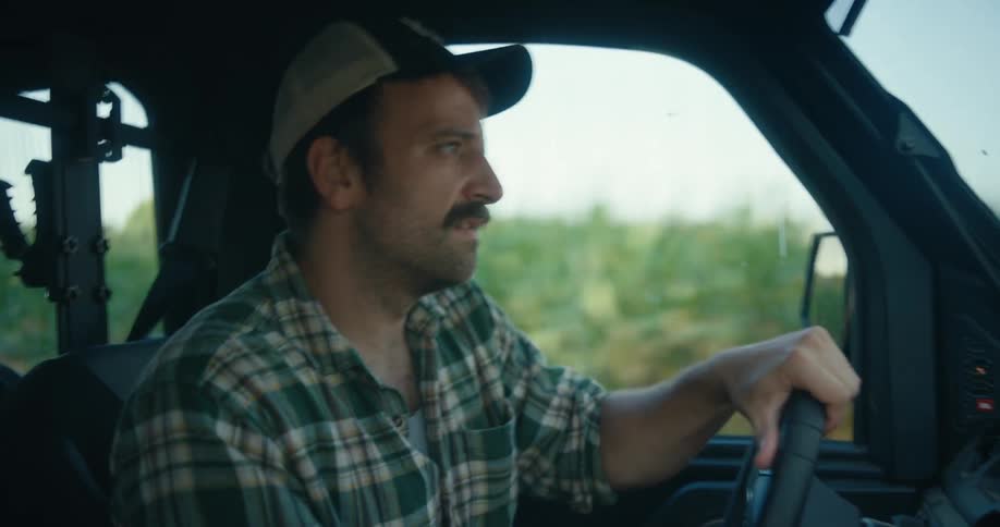 Close up profile of a man with a mustache wearing a plaid shirt and trucker hat driving a utility vehicle through the countryside