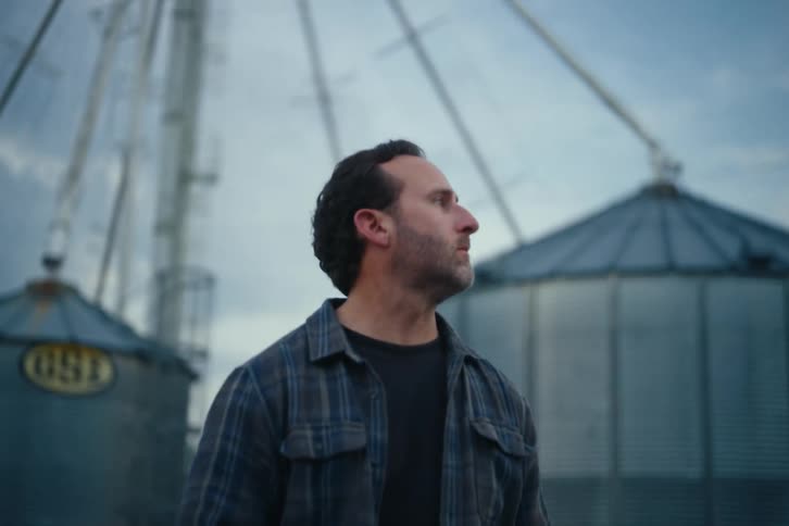 A thoughtful farmer stands in front of large grain silos at dusk, looking out over his land